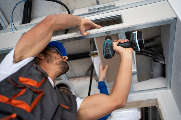Construction worker in hard hat using a drill to install ceiling drywall with a colleague | Al Janah Al Malaky