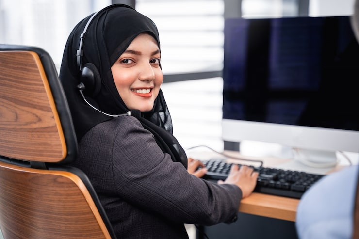 Smiling woman in a headset and black hijab working at a computer in an office | Al Janah Al Malaky