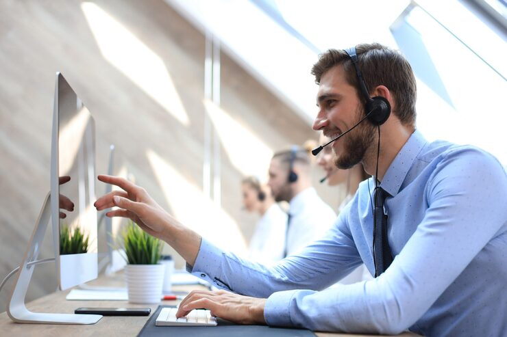 A smiling male call center agent wearing a headset and working at a desk.